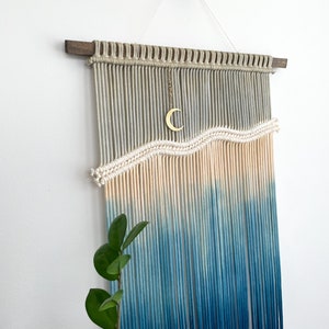 May include: A macrame wall hanging with a wave design in shades of blue and cream. The hanging is made with natural cotton cord and features a crescent moon charm.