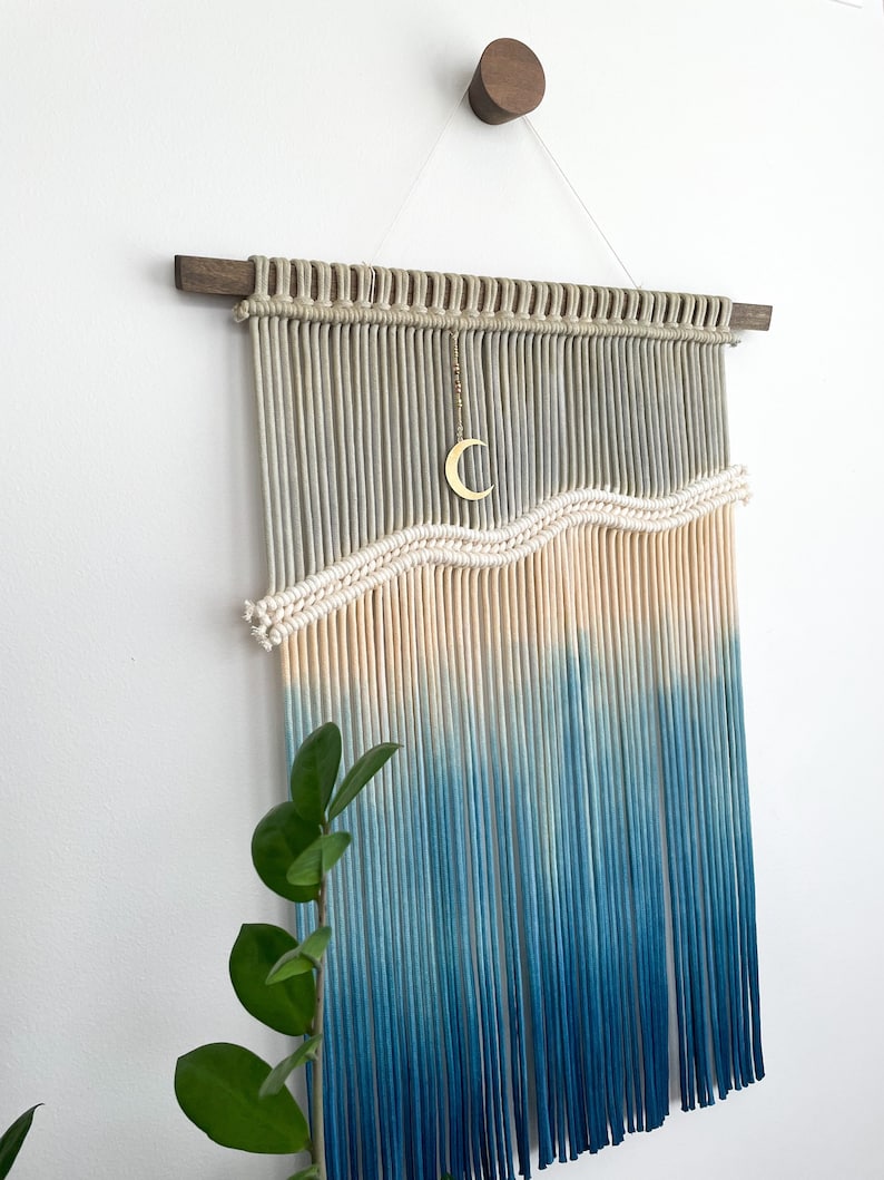 May include: A macrame wall hanging with a wave design in shades of blue and cream. The hanging is made with natural cotton cord and features a crescent moon charm.