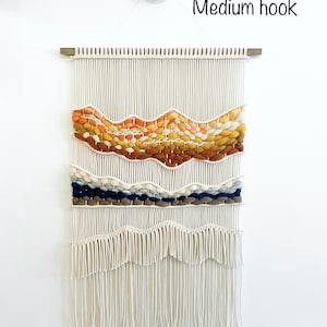 May include: A white macrame wall hanging with a woven design featuring orange, yellow, brown, blue, and white yarn. The hanging is attached to a wooden dowel rod and is hung on a white wall with a medium-sized hook.