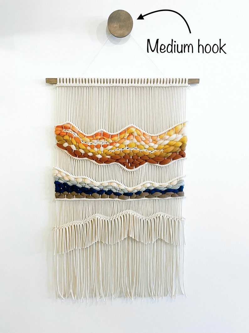 May include: A white macrame wall hanging with a woven design featuring orange, yellow, brown, blue, and white yarn. The hanging is attached to a wooden dowel rod and is hung on a white wall with a medium-sized hook.