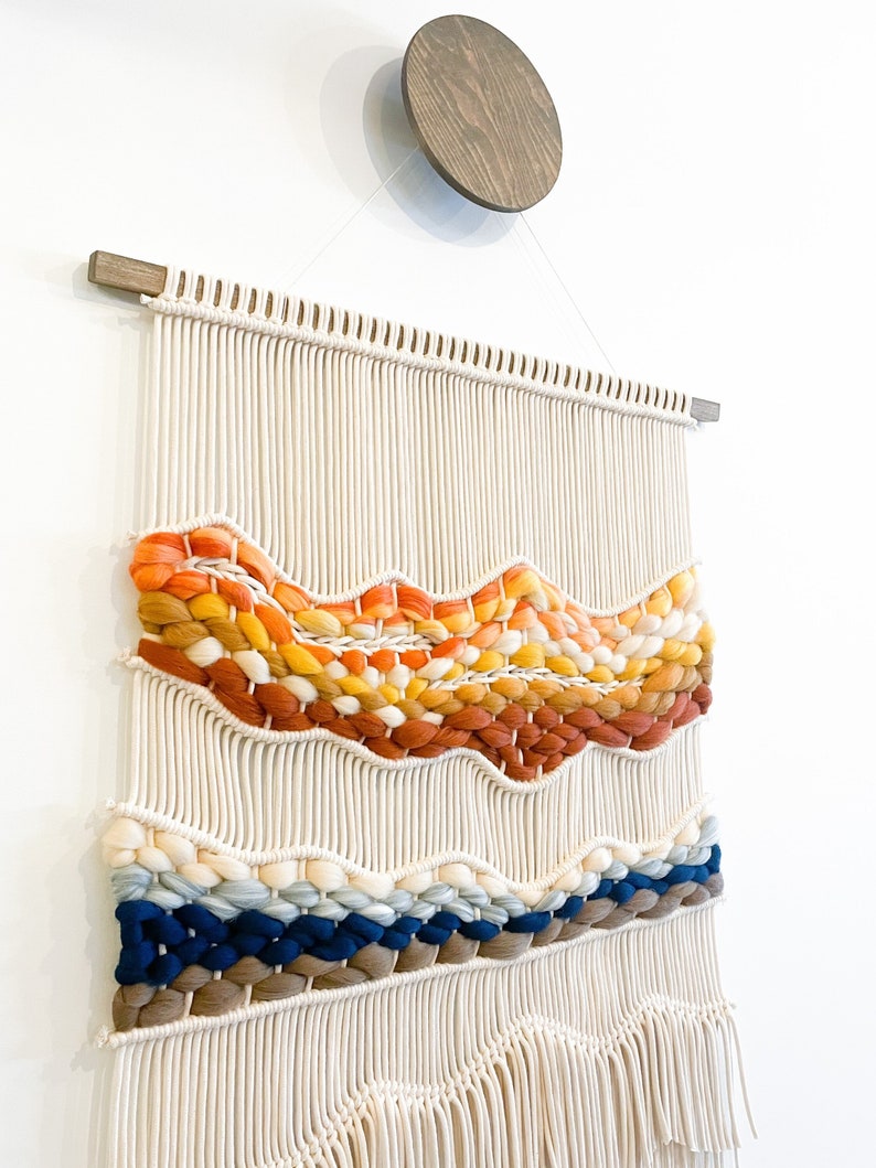 May include: A woven wall hanging with a white background and colorful yarn accents. The hanging features a wavy pattern with orange, yellow, red, blue, and brown yarn. The hanging has a wooden dowel at the top and a fringe at the bottom.