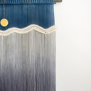 May include: A wall hanging made of macrame with a blue and white ombre design. The hanging features a wavy pattern and a gold circle ornament.