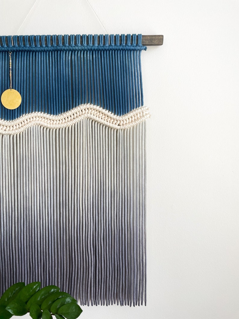 May include: A wall hanging made of macrame with a blue and white ombre design. The hanging features a wavy pattern and a gold circle ornament.