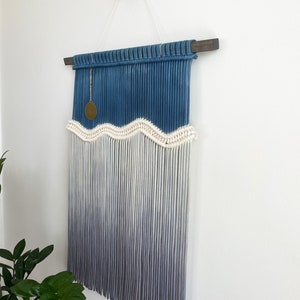 May include: A blue and white macrame wall hanging with a wavy pattern and a gold charm. The hanging is made with cotton cord and has a wooden dowel at the top.