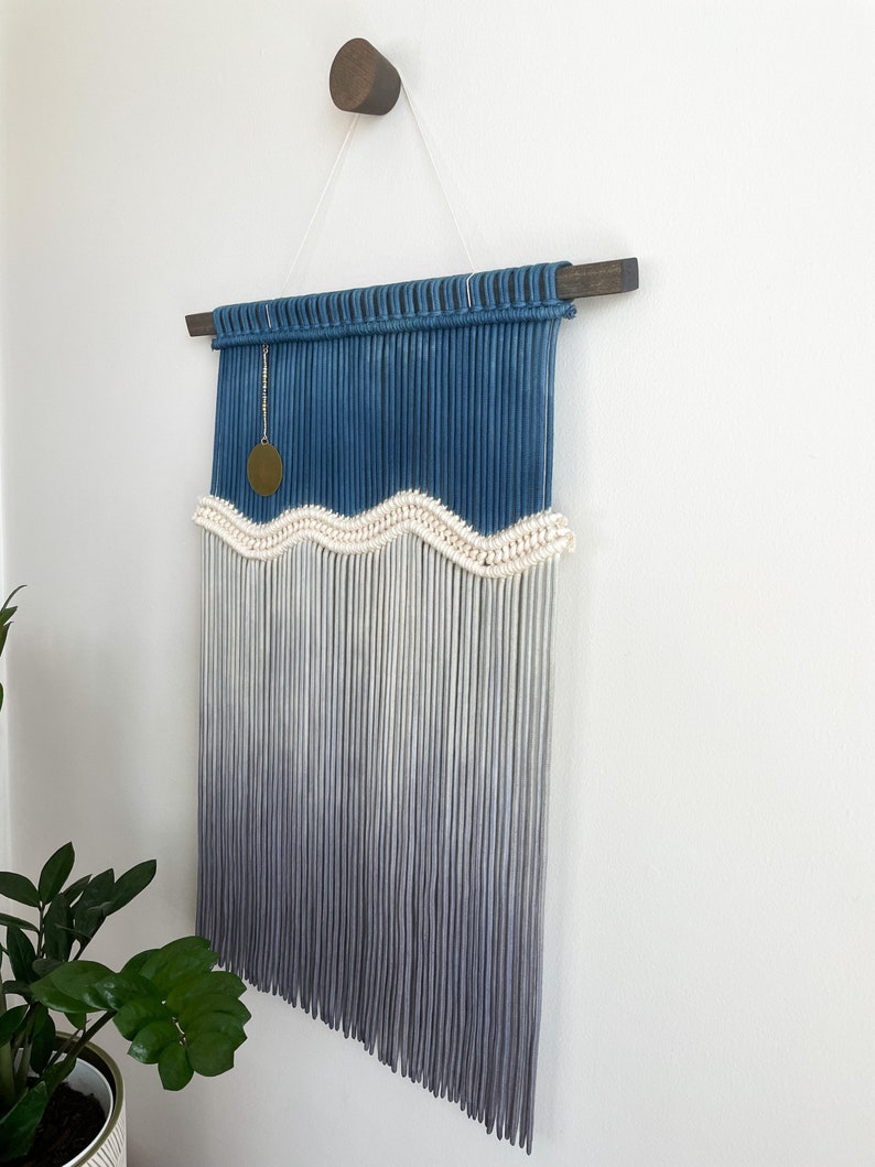 May include: A blue and white macrame wall hanging with a wavy pattern and a gold charm. The hanging is made with cotton cord and has a wooden dowel at the top.