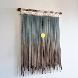 Könnte beinhalten: Ein Wandbehang mit einem Farbverlauf aus blauem und braunem Garn, mit einem weißen gewebten Rand oben. Ein kleiner goldener Kreis hängt in der Mitte des Behangs.