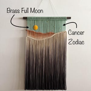 May include: A green, orange, and beige macrame wall hanging with a brass full moon charm. The hanging is labeled "Cancer Zodiac".