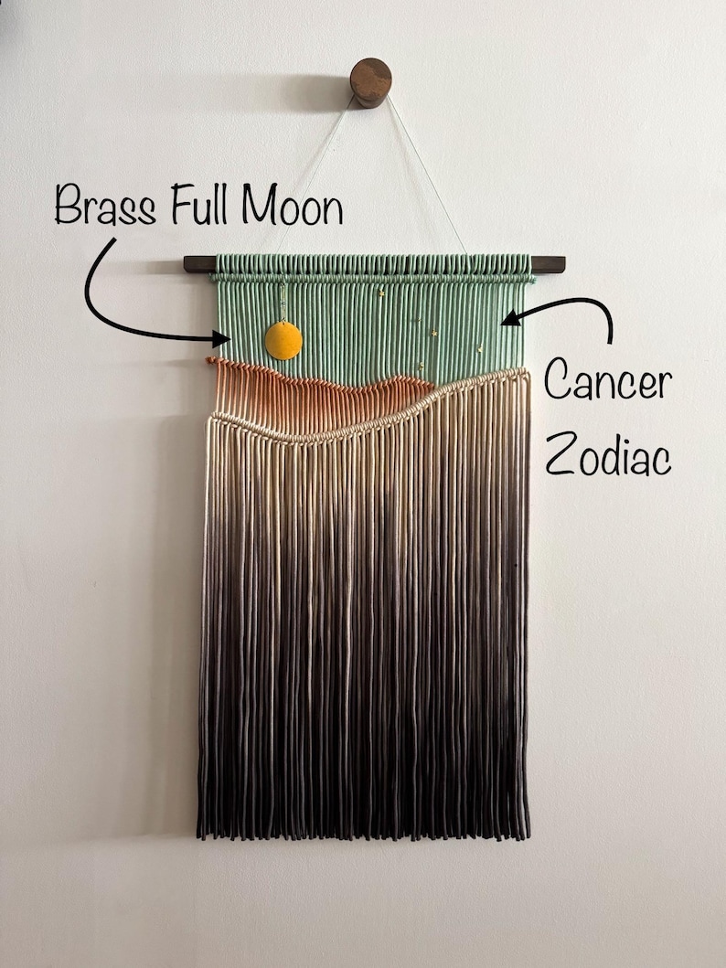 May include: A green, orange, and beige macrame wall hanging with a brass full moon charm. The hanging is labeled "Cancer Zodiac".