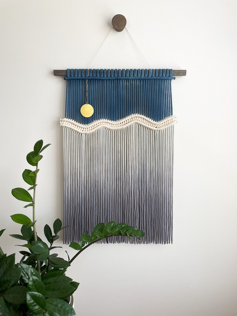 May include: A blue and white macrame wall hanging with a wavy pattern and a gold circle hanging from the top. The wall hanging is made with cotton cord and has a wooden dowel at the top.