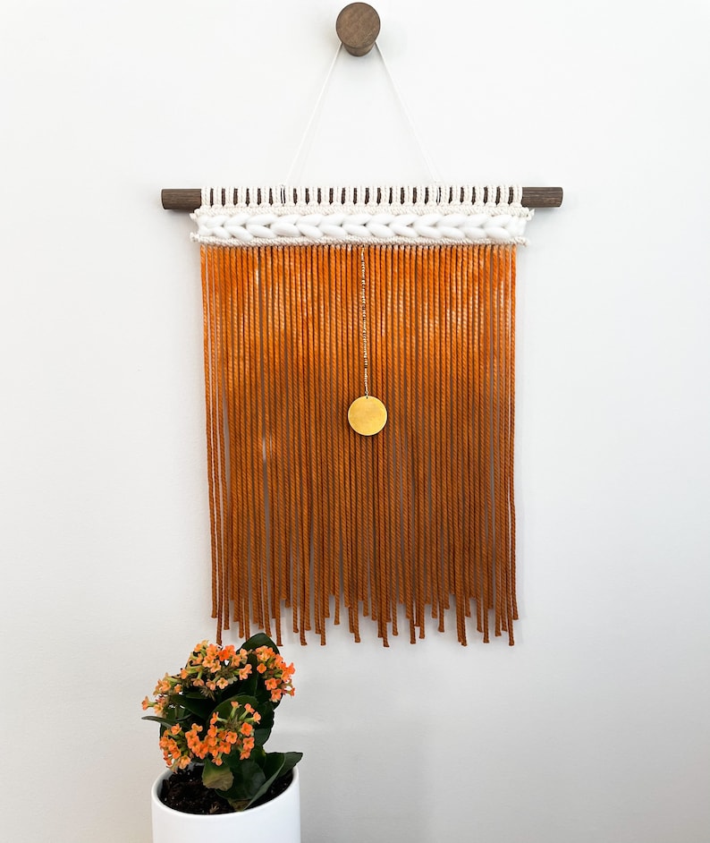May include: A macrame wall hanging with a white and orange ombre fringe and a gold circle pendant. The hanging is attached to a wooden dowel rod.