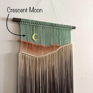 May include: A green, beige, and brown macrame wall hanging with a crescent moon charm. The hanging is made with a variety of cords and has a wooden dowel at the top.
