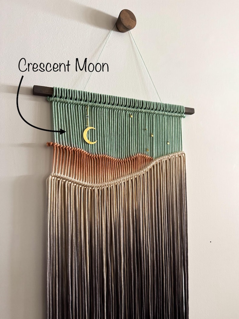 May include: A green, beige, and brown macrame wall hanging with a crescent moon charm. The hanging is made with a variety of cords and has a wooden dowel at the top.