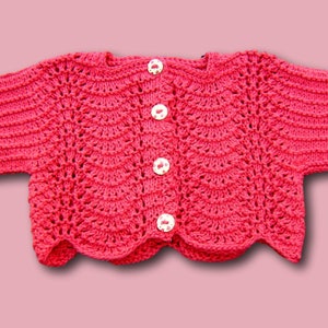 Könnte beinhalten: Eine rosa Strickjacke mit kurzen Ärmeln und Knopfleiste. Die Jacke hat einen gewellten Saum und ist mit einem zarten Strickmuster gearbeitet.