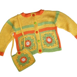 Könnte beinhalten: Eine handgefertigte gelbe Häkeljacke mit grünen und orangefarbenen Streifen und blütenförmigen Taschen. Die Strickjacke hat passende Knöpfe und eine kleine Handtasche mit Blumenmuster. Das Outfit ist aus Garn gefertigt.