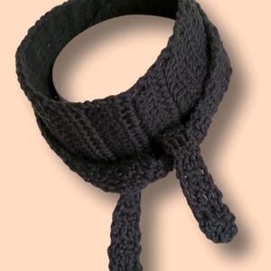 Könnte beinhalten: Ein schwarzer Strick Schal mit langen Bändern. Der Schal ist in der Mitte gefaltet und am Hals gebunden.
