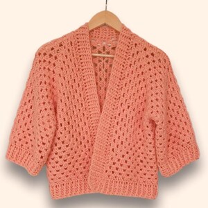 Könnte beinhalten: Ein peachfarbener Häkelcardigan mit einem zarten Lochmuster. Der Cardigan hat einen V-Ausschnitt und kurze Ärmel.