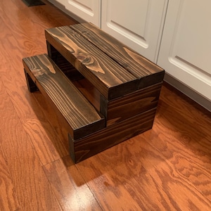 May include: A two-step wooden stool with a dark brown stain. The stool is made of solid wood and has a simple, rustic design.