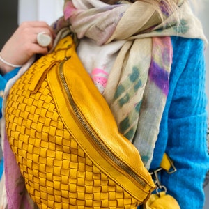 May include: A woven yellow leather fanny pack with a zipper closure. The bag has a small matching coin purse attached. The background includes a blue top, a patterned scarf, and jewelry.