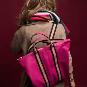 Könnte beinhalten: Eine leuchtend pinkfarbene Tragetasche mit goldenem Reißverschluss und gestreiften Trägern. Die Träger sind marineblau mit einem beigefarbenen Streifen in der Mitte. Die Tasche wird über der Schulter getragen. Ein farbenfroher Schal ist über der Schulter drapiert.