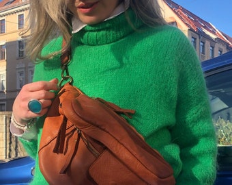 May include: A brown leather fanny pack with a front pocket and zipper closure. The bag is worn over a bright green sweater and white collared shirt. The bag has a worn, vintage look.