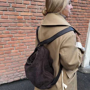 Op de afbeelding: Een donkerbruine suède schoudertas met een zwarte leren riem wordt gedragen over een beige jas. De tas is nonchalant en heeft een zachte textuur. De jas is neutraal van kleur en heeft een klassiek ontwerp.