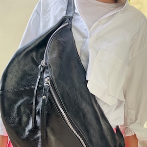 Könnte beinhalten: Eine schwarze Leder-Umhängetasche mit einem silbernen Reißverschluss und einem langen Riemen. Die Tasche wird über der Schulter einer Person getragen, die ein weißes Hemd trägt.
