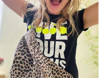 Puede incluir: Una mujer con una camiseta negra con el texto "Live Your Dreams" en letras blancas y amarillas, y una riñonera de estampado de leopardo.