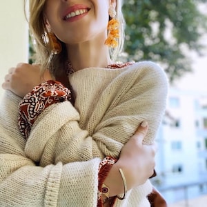 May include: A woman wearing a cream-colored knitted sweater with a brown leather bag over her shoulder. She is wearing a red and white patterned shirt underneath the sweater and has on a gold bracelet with a red stone. She is also wearing orange tassel earrings.