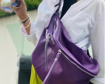 May include: A purple leather crossbody bag with a silver zipper and a long strap. The bag is worn over the shoulder of a person wearing a white shirt and a green skirt.