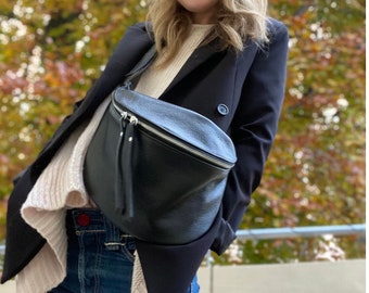 May include: A woman wearing a black blazer, a white sweater, blue jeans, and a black beanie. She is carrying a black leather fanny pack with a silver zipper.