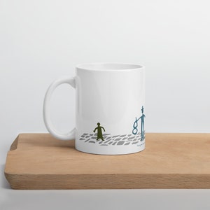 May include: A white ceramic mug featuring a graphic illustration of a small olive green figure and a taller blue figure holding a blue object, both standing on a gray cobblestone path. The design includes small text near the base of the mug.