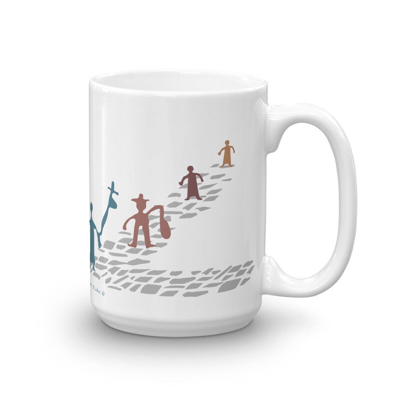 May include: A white ceramic mug featuring a graphic illustration of four stylized figures walking along a gray cobblestone path. The figures are rendered in blue, brown, and tan colors, with one figure carrying a bag and another holding a tall staff.
