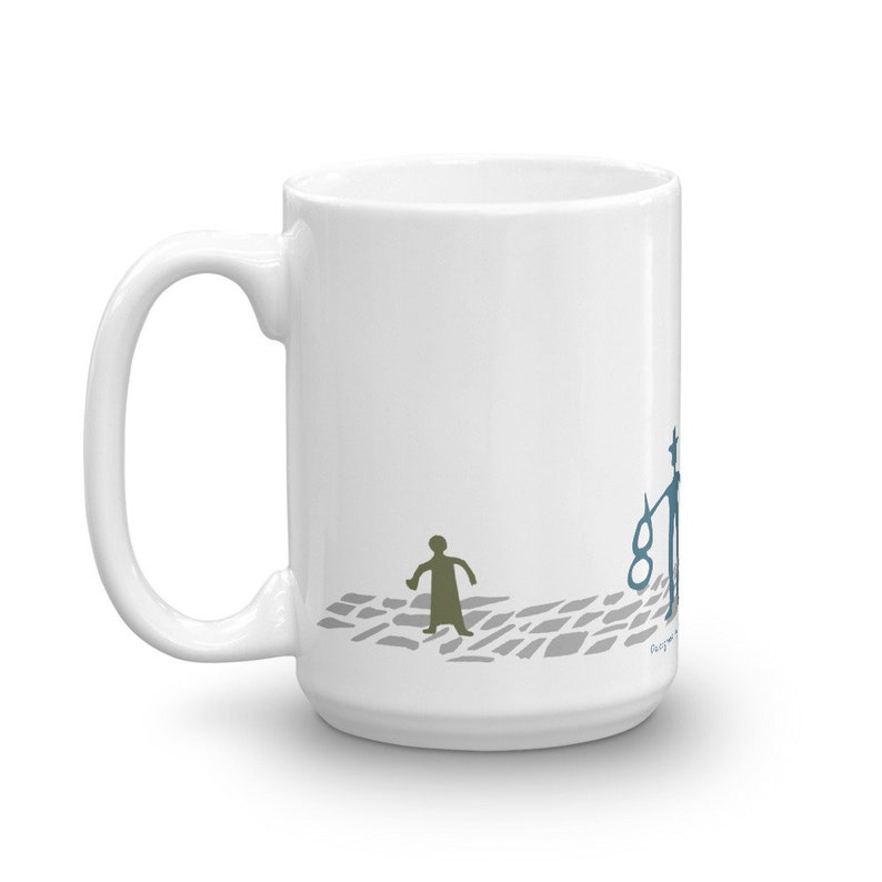 May include: A white ceramic mug featuring a graphic illustration of a small olive-colored figure walking on a gray cobblestone path toward a blue figure holding a circular object. The design includes small text near the base of the blue figure.