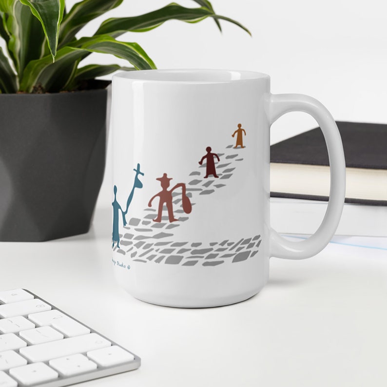 May include: A white ceramic mug featuring a graphic of four stylized figures walking along a gray cobblestone path. The figures are colored in blue, brown, dark red, and orange, with some carrying bags. The design includes small text near the base.