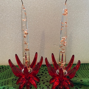 May include: Two decorative candle holders. Each has a clear, cylindrical candle with copper-colored flakes inside. The base is a vibrant red, flower-shaped design. The candles are approximately 10 inches tall.