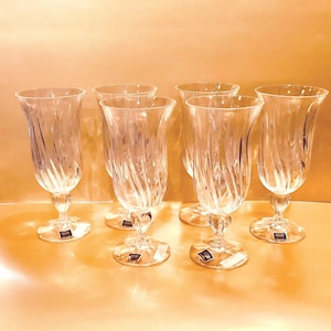 May include: Set of six clear crystal champagne flutes. Each glass features a spiral design on the bowl and a decorative stem. The glasses are displayed on a light-colored surface, showcasing their elegant design.