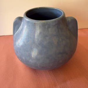May include: A rounded ceramic vase featuring a mottled periwinkle blue glaze and two integrated side handles. The vessel has a wide, circular opening at the top and a smooth, bulbous body with subtle variations in the blue color throughout the surface.