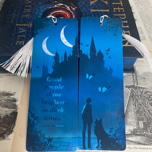 Peut inclure: Deux marque-pages bleus avec des pompons blancs. L'un présente un croissant de lune, une silhouette de château et le texte "Good people shine brighter in dark times". L'autre montre une silhouette de personne et de chien avec des papillons.