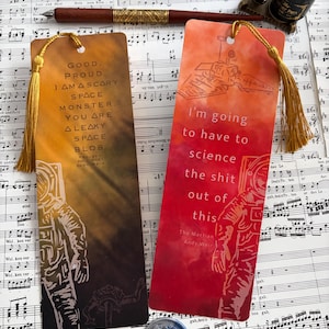 Puede incluir: Dos marcapáginas rectangulares con borlas doradas. Uno es naranja y marrón con una ilustración de astronauta y texto. El otro es rojo y naranja con una ilustración de rover espacial y texto. Ambos están sobre un fondo de partituras.