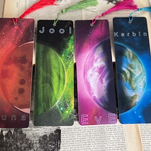 Peut inclure: Quatre marque-pages colorés avec des pompons. Chaque marque-page présente un design céleste avec une planète et un nom : Duna, Jool, Eve et Korbin. Les marque-pages sont rectangulaires et en matériau brillant.