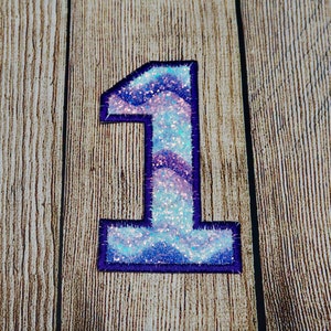 May include: A sparkly blue and purple number one with a dark blue outline. The number is on a wood background.