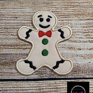 May include: Embroidered gingerbread man patch with a red bow tie, black accents, and green buttons. The patch has a cream-coloured background and a stitched border. The patch is on a wooden surface.