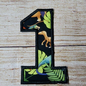 May include: Embroidered number one patch with a black border. The interior features a black background with a dinosaur and tropical leaf pattern. The patch is set against a light wood grain background.