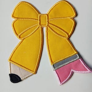 May include: A yellow fabric bow-shaped patch with a pencil design. The pencil has a gray band, a pink eraser, and a black tip. The patch is likely for decorating clothing or accessories.