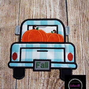 以下が含まれることがあります： 青い白のチェック柄のピックアップトラックが、荷台を開けてオレンジ色のカボチャを3つ運んでいます。トラックのナンバープレートには「Fall」と書いてあります。