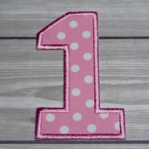 May include: Pink felt number one with white polka dots. The number is outlined in a darker pink.