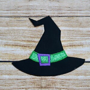 May include: A black witch hat with a green glitter band and a purple buckle. The hat is set against a light brown wooden background. The hat's brim is wide and the crown is pointed.
