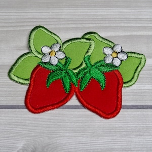 May include: Two red embroidered strawberries with green leaves and white flowers. The strawberries are sewn onto a light brown fabric.