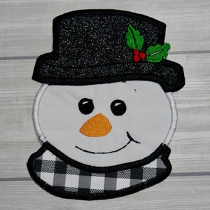 Könnte beinhalten: Ein weißes Schneemann-Gesicht mit schwarzen Augen, einer orangefarbenen Nase und einem schwarz-weiß karierten Schal. Der Schneemann trägt einen schwarzen Zylinder mit einem grünen Stechpalmenzweig.