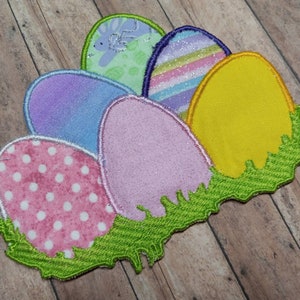 May include: A set of five embroidered felt Easter eggs in a variety of colors and patterns, including pink with white polka dots, purple, light blue, yellow, and a multi-colored striped egg. The eggs are arranged in a semi-circle and are surrounded by green felt grass.